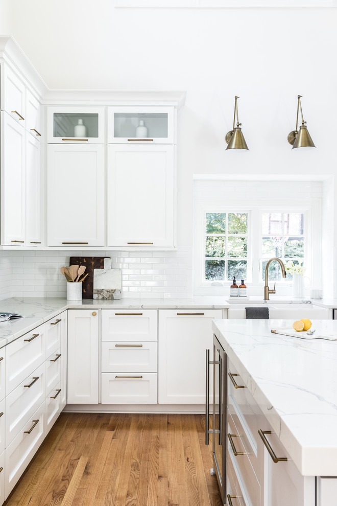 Inspiration for a large traditional l-shaped kitchen in Charlotte with a belfast sink, shaker cabinets, white cabinets, engineered stone countertops, white splashback, an island, metro tiled splashback and light hardwood flooring.