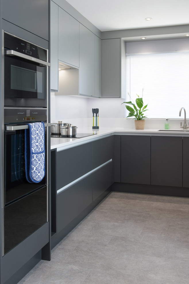 This is an example of a small modern u-shaped enclosed kitchen in Cornwall with flat-panel cabinets, grey cabinets, composite countertops, white splashback, black appliances, no island and white worktops.