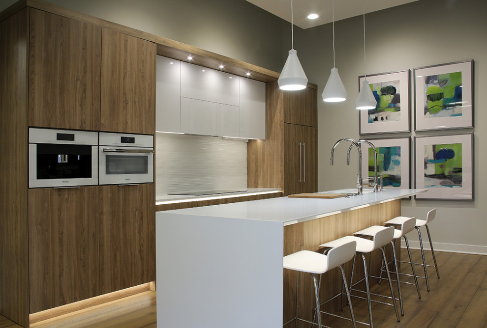 Large modern single-wall open plan kitchen in Grand Rapids with a submerged sink, flat-panel cabinets, light wood cabinets, engineered stone countertops, white splashback, porcelain splashback, integrated appliances, medium hardwood flooring, an island, brown floors and white worktops.
