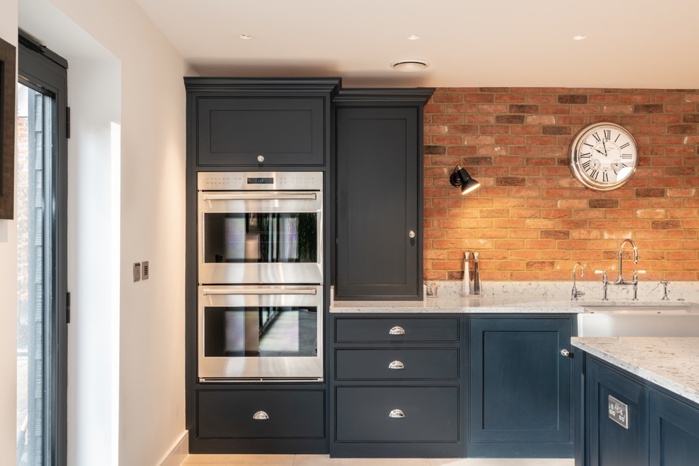 Inspiration for an expansive contemporary single-wall open plan kitchen in Essex with a belfast sink, beaded cabinets, blue cabinets, quartz worktops, multi-coloured splashback, brick splashback, stainless steel appliances, terracotta flooring, an island, beige floors and white worktops.