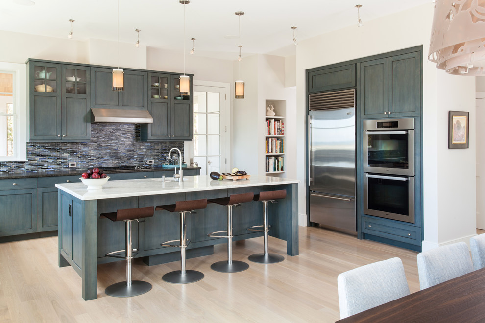 Foto på ett stort maritimt kök, med en undermonterad diskho, luckor med upphöjd panel, blå skåp, marmorbänkskiva, blått stänkskydd, stänkskydd i glaskakel, rostfria vitvaror, ljust trägolv och en köksö