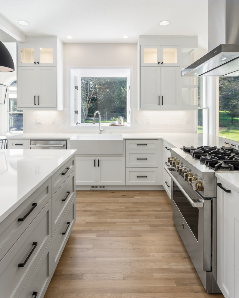 This is an example of a classic l-shaped kitchen/diner in Portland with shaker cabinets, grey cabinets, white splashback, stainless steel appliances, medium hardwood flooring, an island, white worktops, a belfast sink, engineered stone countertops and ceramic splashback.