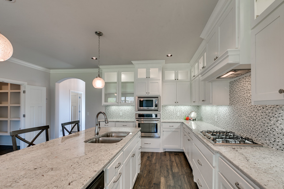 Medium sized traditional l-shaped kitchen/diner in Oklahoma City with a double-bowl sink, shaker cabinets, white cabinets, granite worktops, white splashback, glass tiled splashback, stainless steel appliances, dark hardwood flooring and an island.