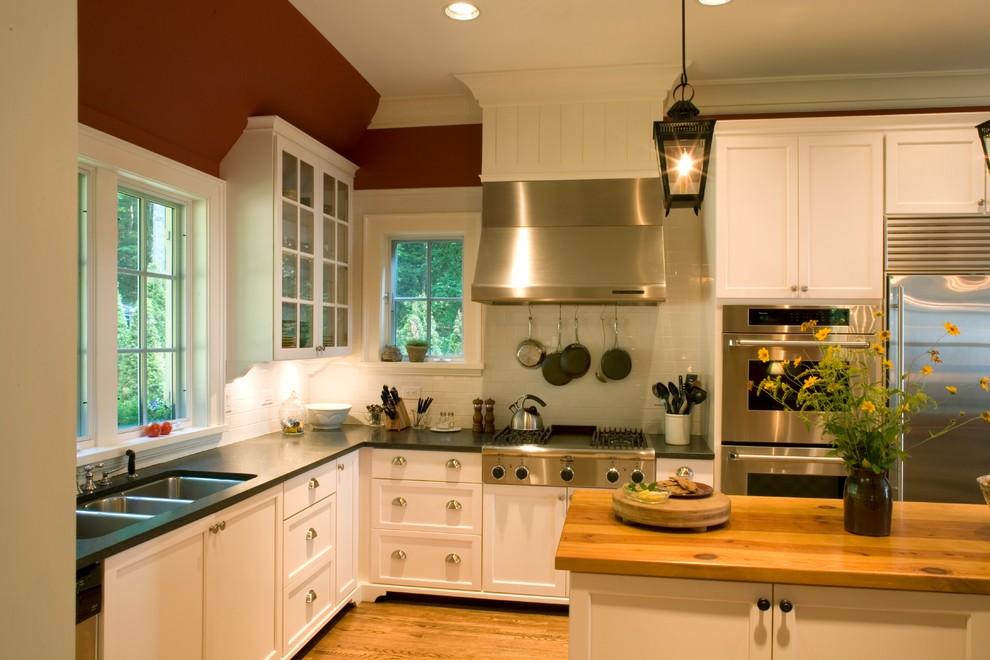 Elegant l-shaped light wood floor enclosed kitchen photo in Charlotte with a triple-bowl sink, shaker cabinets, white cabinets, soapstone countertops, white backsplash, ceramic backsplash, stainless steel appliances and an island