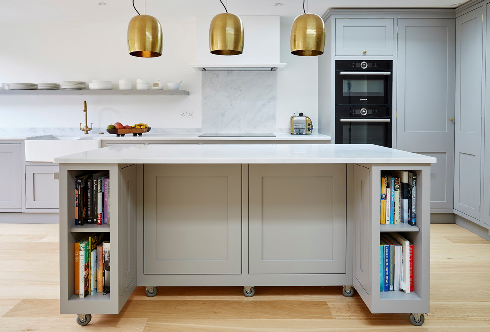 This is an example of a medium sized contemporary l-shaped kitchen/diner in London with a belfast sink, shaker cabinets, grey cabinets, composite countertops, white splashback, black appliances, medium hardwood flooring and an island.