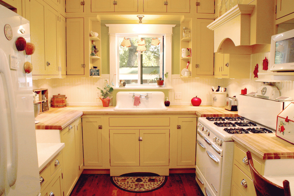 Foto på ett avskilt, litet lantligt u-kök, med gula skåp, träbänkskiva, vitt stänkskydd, vita vitvaror, mörkt trägolv, en nedsänkt diskho och skåp i shakerstil