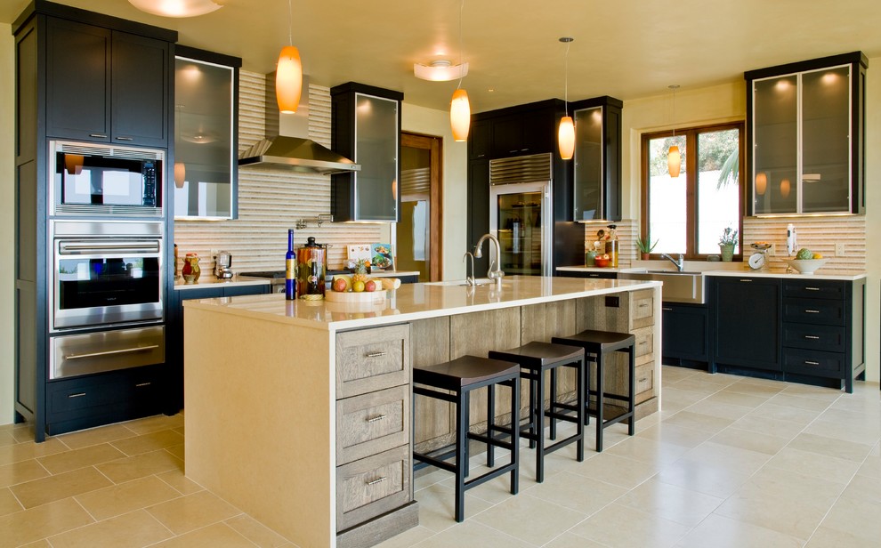 Photo of a medium sized world-inspired single-wall kitchen/diner in Santa Barbara with a double-bowl sink, shaker cabinets, black cabinets, beige splashback, stainless steel appliances, travertine flooring, an island, brown floors and beige worktops.
