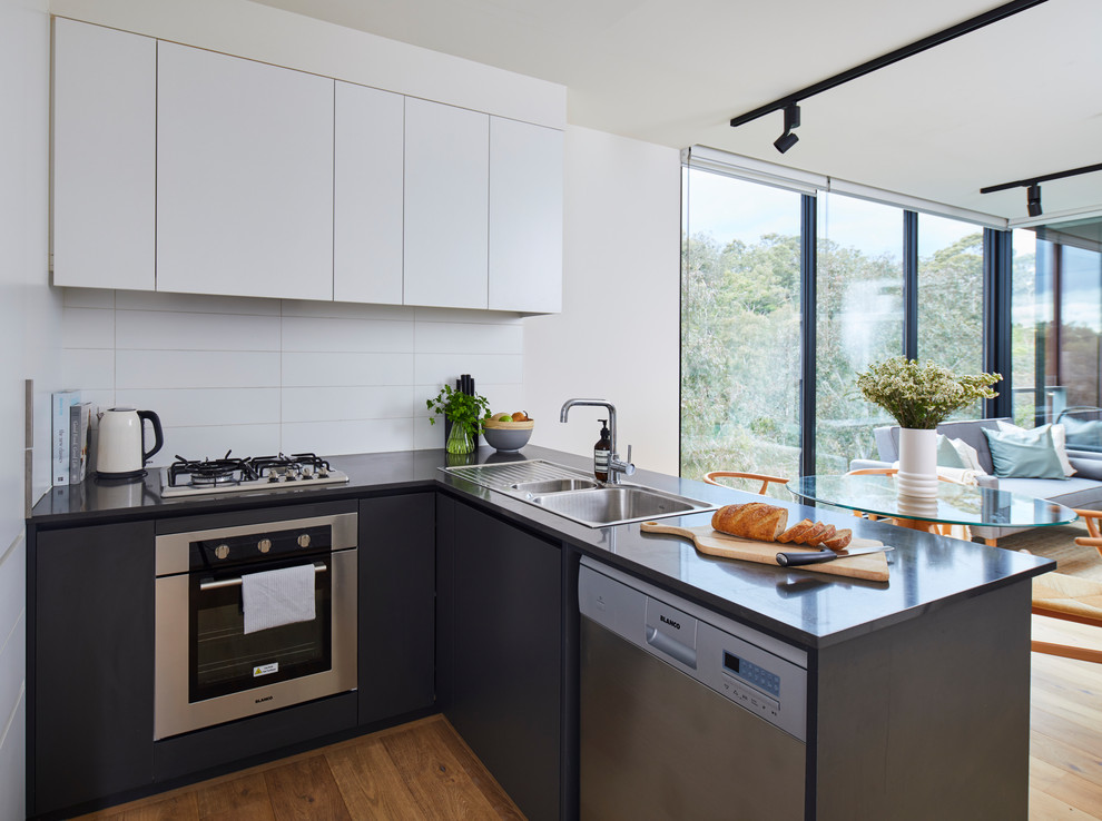 Inspiration for a contemporary l-shaped kitchen in Melbourne with a built-in sink, flat-panel cabinets, black cabinets, white splashback, stainless steel appliances, light hardwood flooring, a breakfast bar and black worktops.