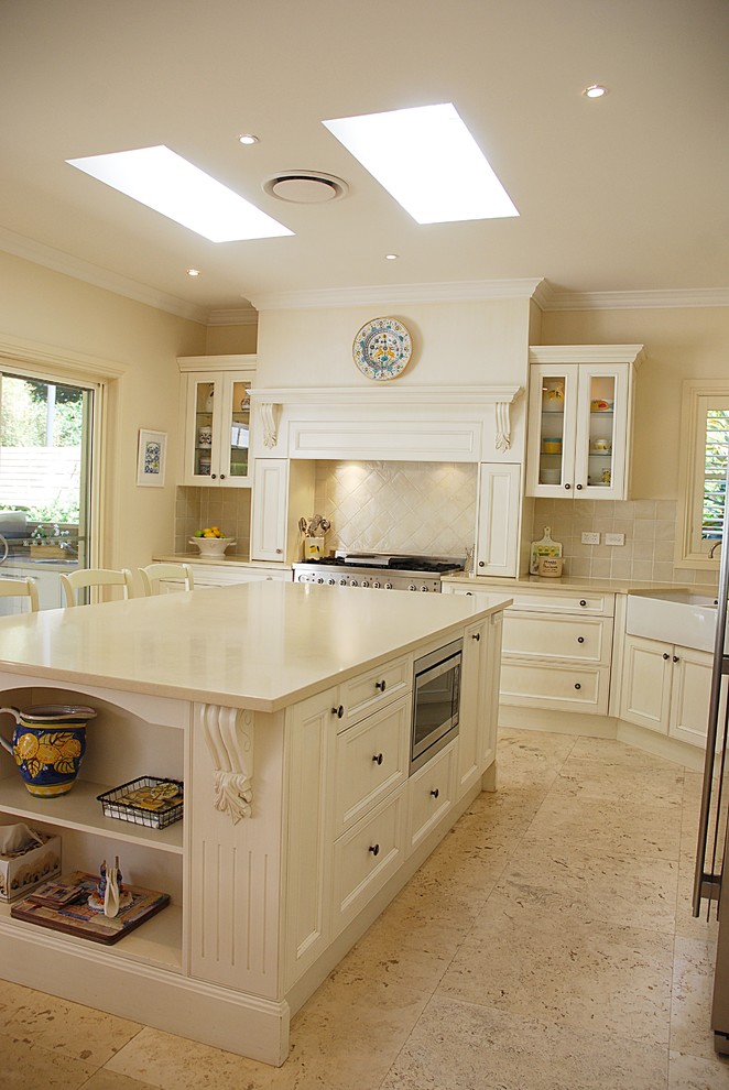 Photo of a large classic l-shaped kitchen/diner in Sydney with a belfast sink, shaker cabinets, white cabinets, engineered stone countertops, beige splashback, ceramic splashback, stainless steel appliances, travertine flooring and an island.