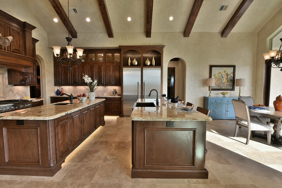 Example of a large tuscan galley travertine floor and beige floor open concept kitchen design in Houston with a farmhouse sink, raised-panel cabinets, brown cabinets, granite countertops, beige backsplash, stone tile backsplash, stainless steel appliances and two islands