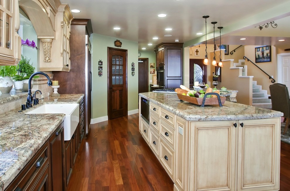 Example of a large tuscan l-shaped medium tone wood floor and brown floor eat-in kitchen design in San Diego with a farmhouse sink, granite countertops, beige backsplash, stone tile backsplash, stainless steel appliances, beaded inset cabinets, beige cabinets and two islands
