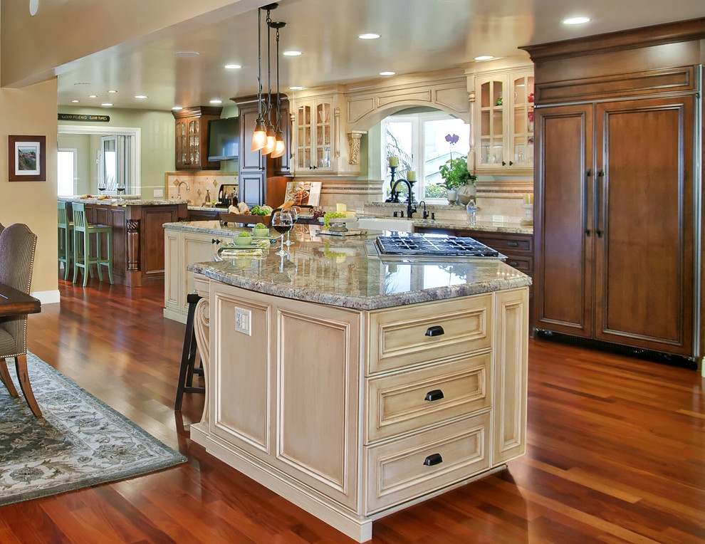 Example of a large tuscan l-shaped medium tone wood floor and brown floor eat-in kitchen design in San Diego with a farmhouse sink, beige cabinets, granite countertops, beige backsplash, stone tile backsplash, stainless steel appliances, beaded inset cabinets and two islands