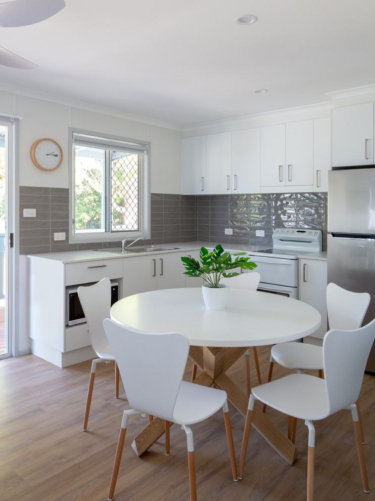 Photo of a small coastal l-shaped open plan kitchen in Gold Coast - Tweed with a single-bowl sink, white cabinets, laminate countertops, grey splashback, ceramic splashback, stainless steel appliances, laminate floors, no island, brown floors and white worktops.