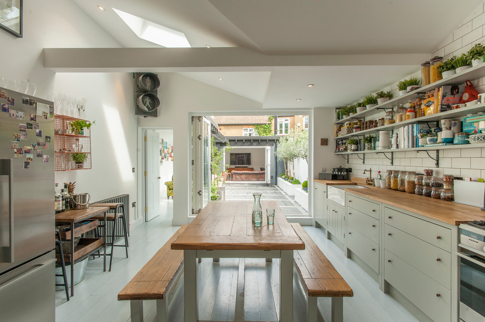 Example of a cottage light wood floor kitchen design in London with a farmhouse sink, flat-panel cabinets, green cabinets, white backsplash, subway tile backsplash and stainless steel appliances