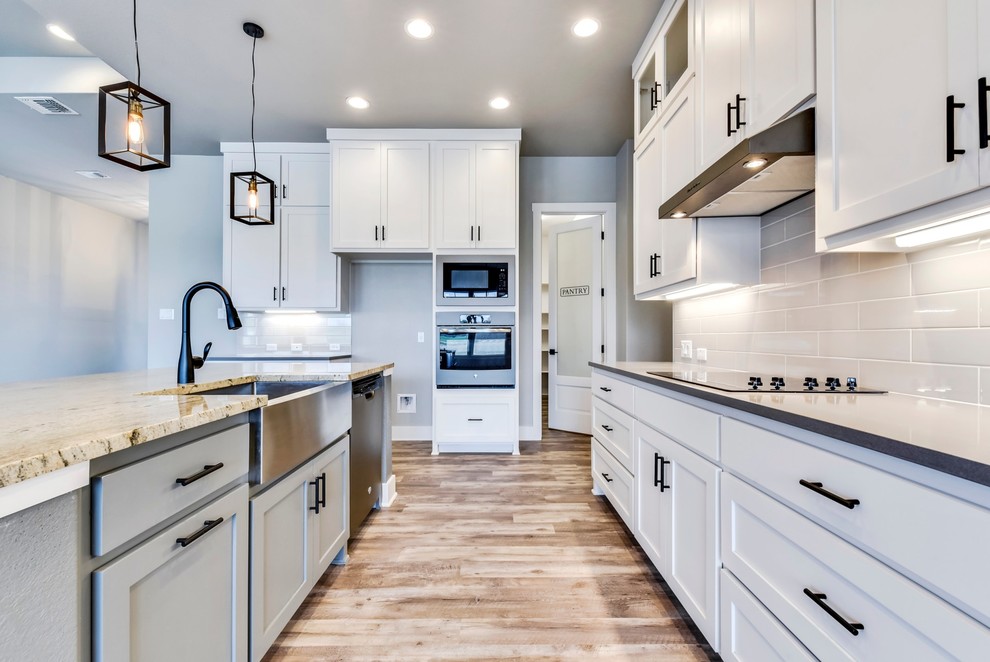Inspiration for a mid-sized transitional l-shaped light wood floor and brown floor open concept kitchen remodel in Austin with a farmhouse sink, recessed-panel cabinets, white cabinets, granite countertops, gray backsplash, subway tile backsplash, stainless steel appliances, an island and gray countertops