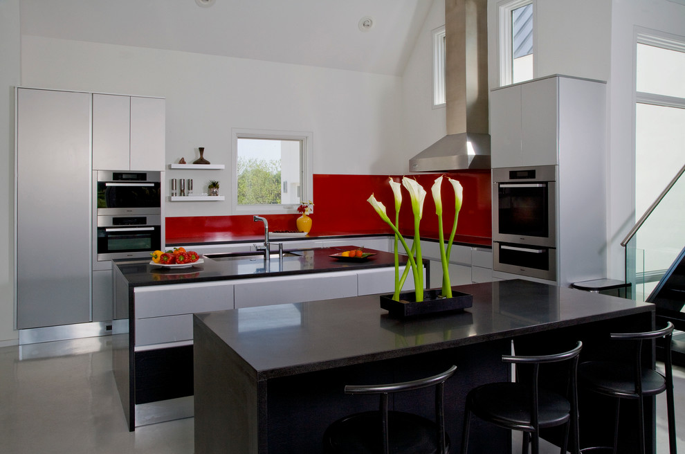 Immagine di una grande cucina design con lavello sottopiano, ante lisce, ante in acciaio inossidabile, top in granito, paraspruzzi rosso, elettrodomestici da incasso, pavimento in cemento e 2 o più isole