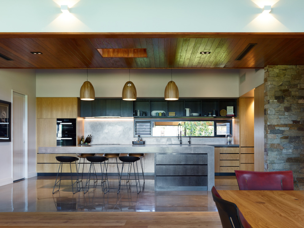 Example of a trendy galley concrete floor, brown floor and wood ceiling open concept kitchen design in Sunshine Coast with flat-panel cabinets, medium tone wood cabinets, black appliances, an island and gray countertops