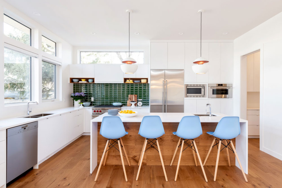 Design ideas for a retro l-shaped open plan kitchen in Other with a submerged sink, flat-panel cabinets, white cabinets, engineered stone countertops, green splashback, metro tiled splashback, stainless steel appliances, medium hardwood flooring, an island, brown floors and white worktops.