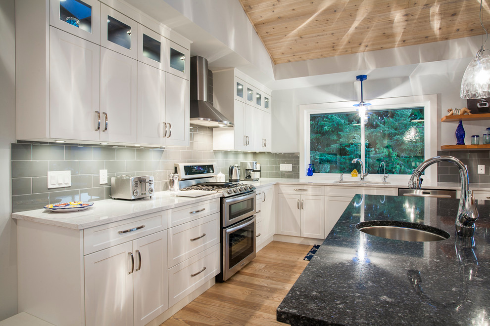 This is an example of a large coastal u-shaped kitchen/diner in Vancouver with a submerged sink, shaker cabinets, white cabinets, granite worktops, grey splashback, glass sheet splashback, stainless steel appliances, light hardwood flooring, an island and brown floors.