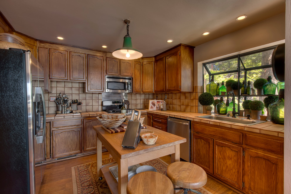Photo of a rustic l-shaped kitchen in Sacramento with a submerged sink, recessed-panel cabinets, dark wood cabinets, tile countertops, beige splashback, ceramic splashback, stainless steel appliances, medium hardwood flooring and an island.