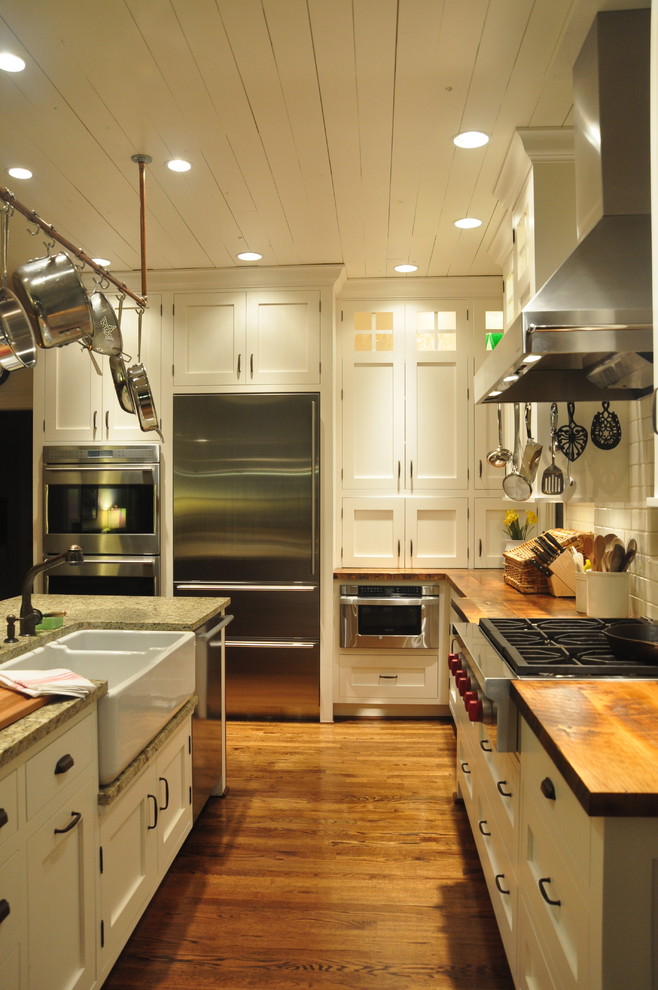 Example of a mid-sized country l-shaped medium tone wood floor kitchen design in Louisville with a farmhouse sink, stainless steel appliances, shaker cabinets, wood countertops, white cabinets, white backsplash, ceramic backsplash and an island