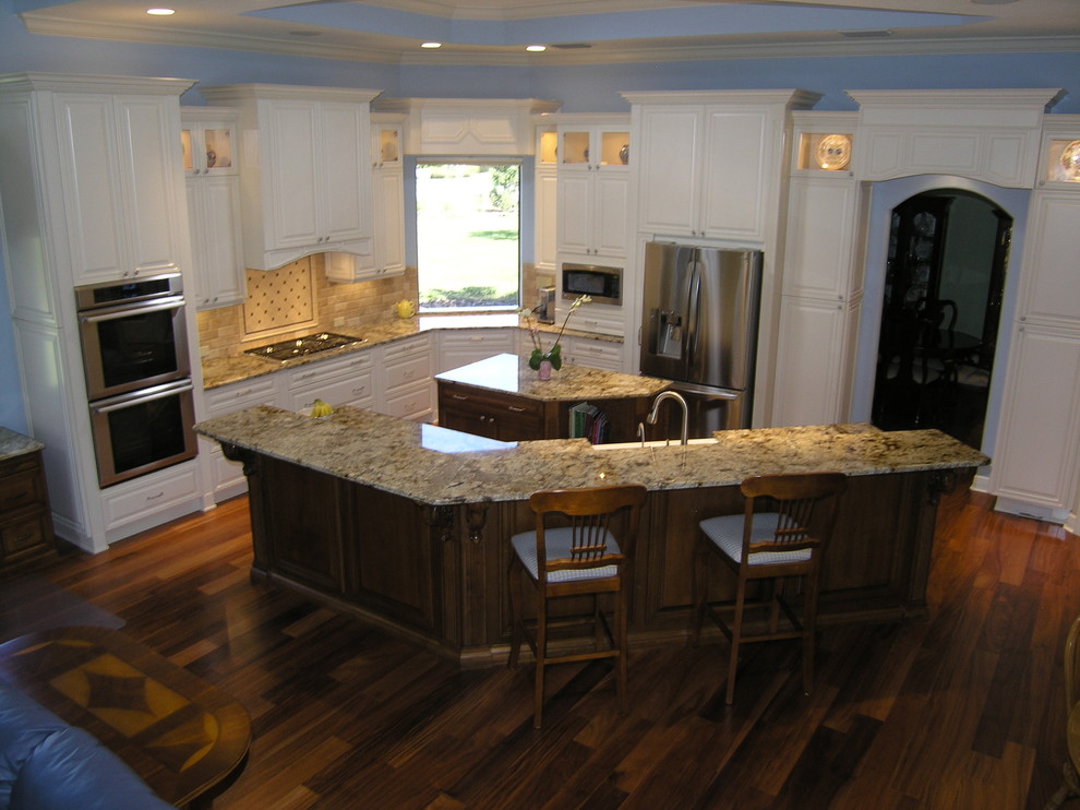 Example of a large classic l-shaped dark wood floor open concept kitchen design in Tampa with a farmhouse sink, raised-panel cabinets, white cabinets, granite countertops, beige backsplash, stone tile backsplash, stainless steel appliances and two islands