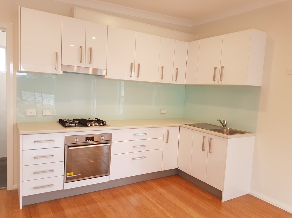 This is an example of a small modern l-shaped open plan kitchen in Sydney with a single-bowl sink, flat-panel cabinets, white cabinets, laminate countertops, green splashback, glass sheet splashback, stainless steel appliances, bamboo flooring and no island.