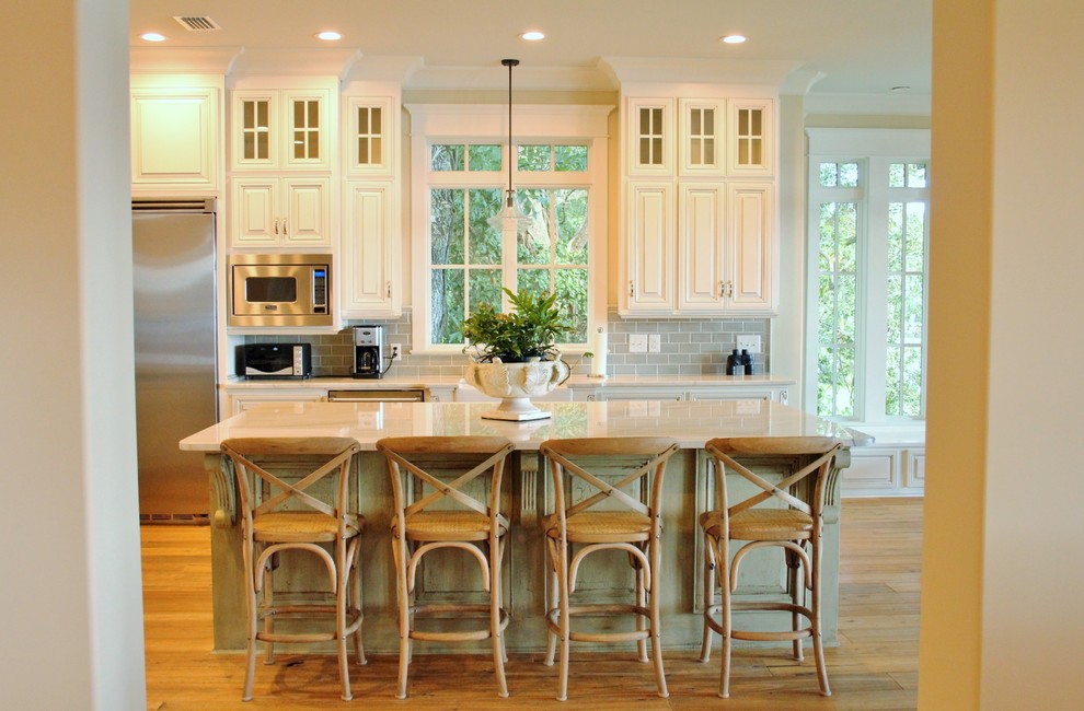 Inredning av ett kök, med en rustik diskho, luckor med upphöjd panel, vita skåp, marmorbänkskiva, vitt stänkskydd, stänkskydd i porslinskakel, rostfria vitvaror, ljust trägolv och en köksö