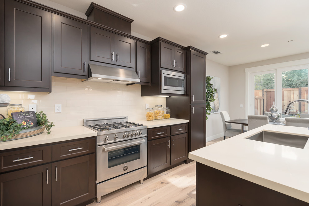 This is an example of a traditional l-shaped kitchen/diner in San Francisco with a belfast sink, shaker cabinets, brown cabinets, engineered stone countertops, beige splashback, ceramic splashback, stainless steel appliances, light hardwood flooring and an island.