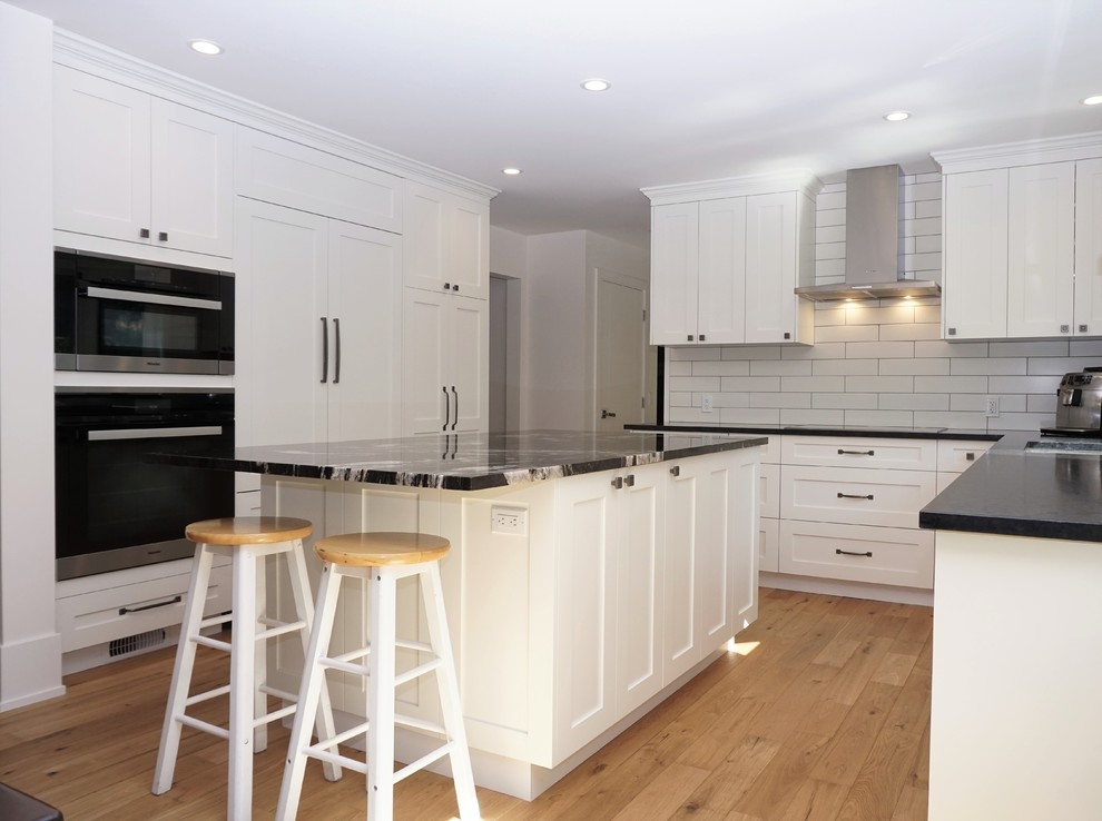 Design ideas for a medium sized traditional kitchen/diner in Calgary with a submerged sink, shaker cabinets, white cabinets, granite worktops, white splashback, light hardwood flooring, an island and black worktops.