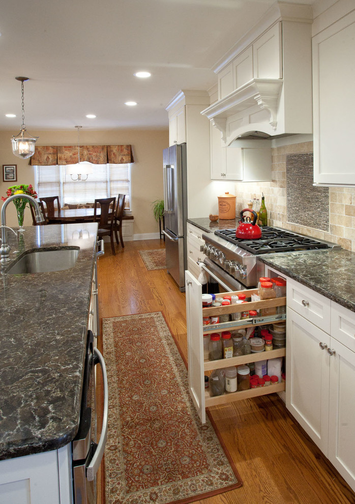 Klassisk inredning av ett kök, med en nedsänkt diskho, luckor med upphöjd panel, vita skåp, granitbänkskiva, grått stänkskydd, stänkskydd i stenkakel, rostfria vitvaror, ljust trägolv och en köksö