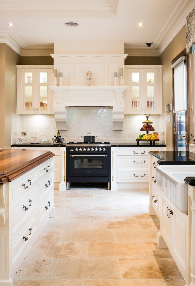 This is an example of a large farmhouse l-shaped enclosed kitchen in Melbourne with a belfast sink, beaded cabinets, white cabinets, engineered stone countertops, white splashback, ceramic splashback, black appliances, travertine flooring and an island.
