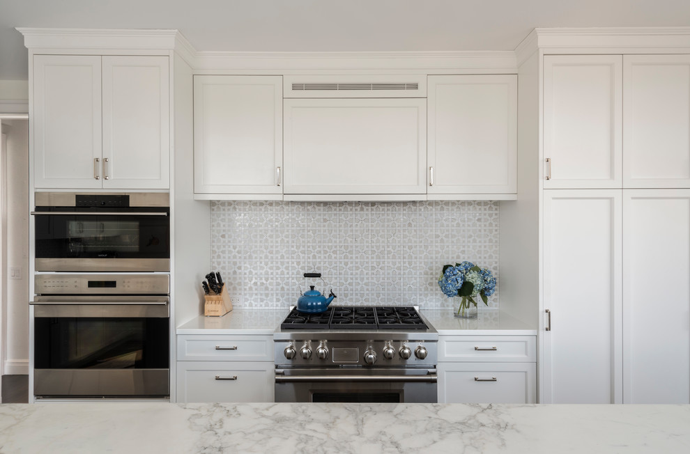 Large traditional l-shaped kitchen/diner in New York with recessed-panel cabinets, white cabinets, glass worktops, white splashback, ceramic splashback, stainless steel appliances and an island.