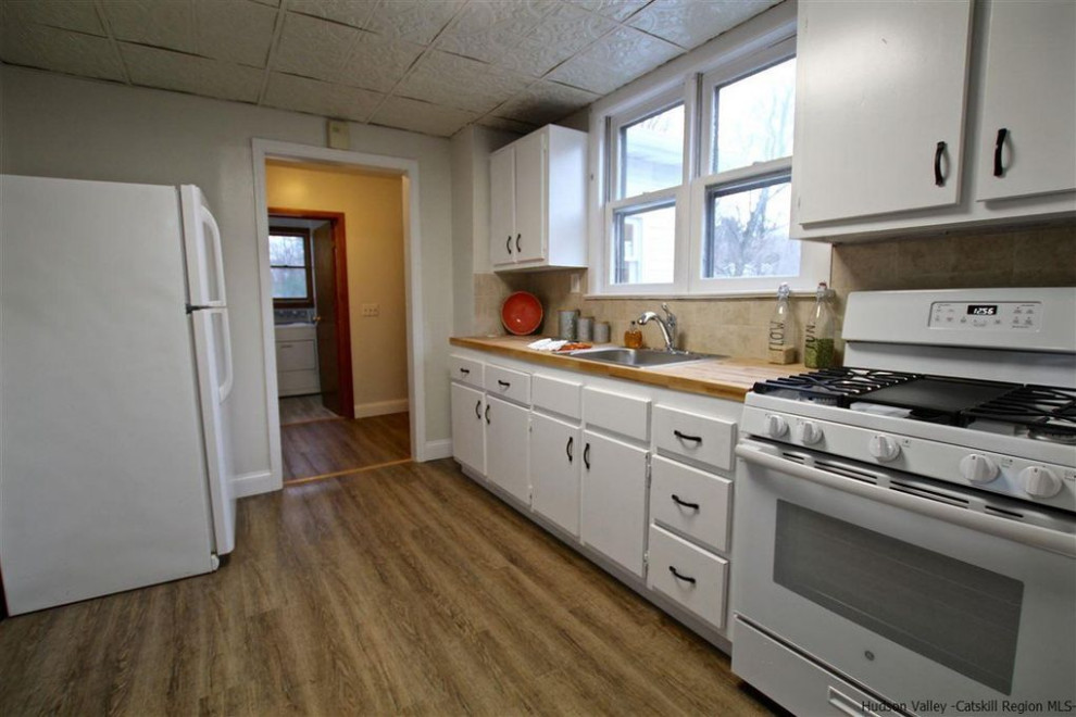 ニューヨークにある小さなラスティックスタイルのおしゃれなキッチン (ドロップインシンク、フラットパネル扉のキャビネット、白いキャビネット、木材カウンター、ベージュキッチンパネル、セラミックタイルのキッチンパネル、白い調理設備、無垢フローリング、アイランドなし、茶色い床、ベージュのキッチンカウンター) の写真