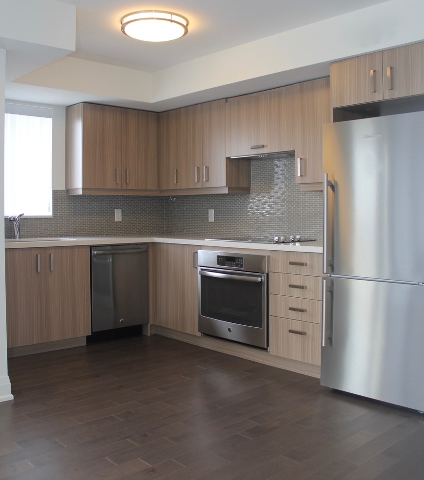 This is an example of a medium sized contemporary l-shaped open plan kitchen in Toronto with flat-panel cabinets, a submerged sink, light wood cabinets, engineered stone countertops, grey splashback, glass tiled splashback, stainless steel appliances and vinyl flooring.