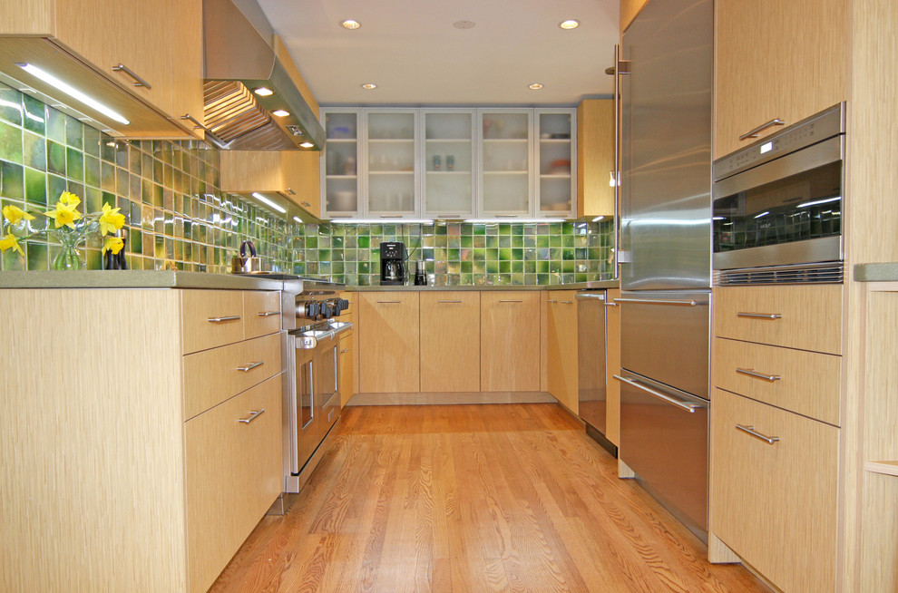 Cette photo montre une cuisine tendance en U et bois clair avec un électroménager en acier inoxydable, un placard à porte plane et une crédence verte.