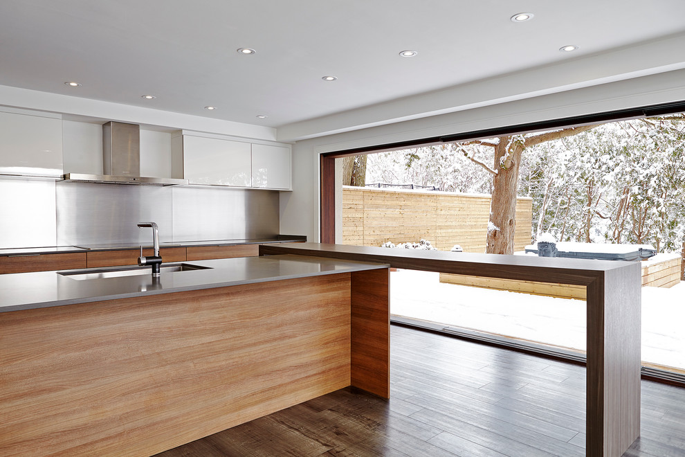 This is an example of a midcentury galley open plan kitchen in Toronto with a submerged sink, flat-panel cabinets, medium wood cabinets, engineered stone countertops, metallic splashback, metal splashback, integrated appliances, vinyl flooring and a breakfast bar.