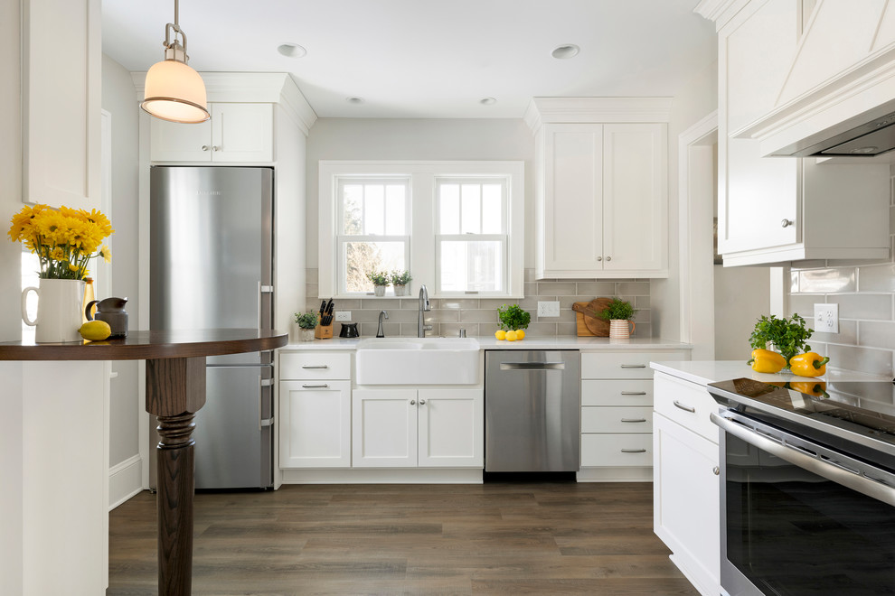 Design ideas for a small farmhouse l-shaped kitchen in Minneapolis with a belfast sink, shaker cabinets, white cabinets, grey splashback, metro tiled splashback, stainless steel appliances, medium hardwood flooring, a breakfast bar, brown floors and white worktops.