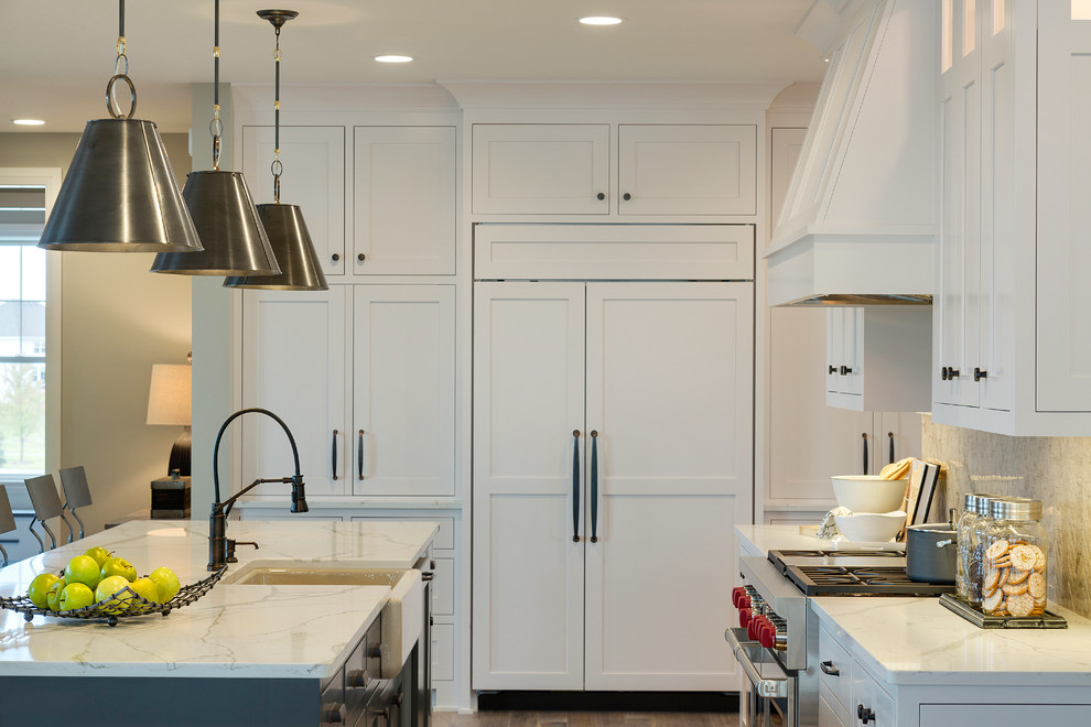 Photo of a large classic l-shaped kitchen in Minneapolis with a belfast sink, white cabinets, engineered stone countertops, grey splashback, ceramic splashback, integrated appliances, medium hardwood flooring and an island.