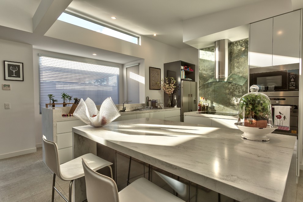 Modern inredning av ett mellanstort kök, med en undermonterad diskho, släta luckor, vita skåp, marmorbänkskiva, stänkskydd med metallisk yta, glaspanel som stänkskydd, rostfria vitvaror och en köksö