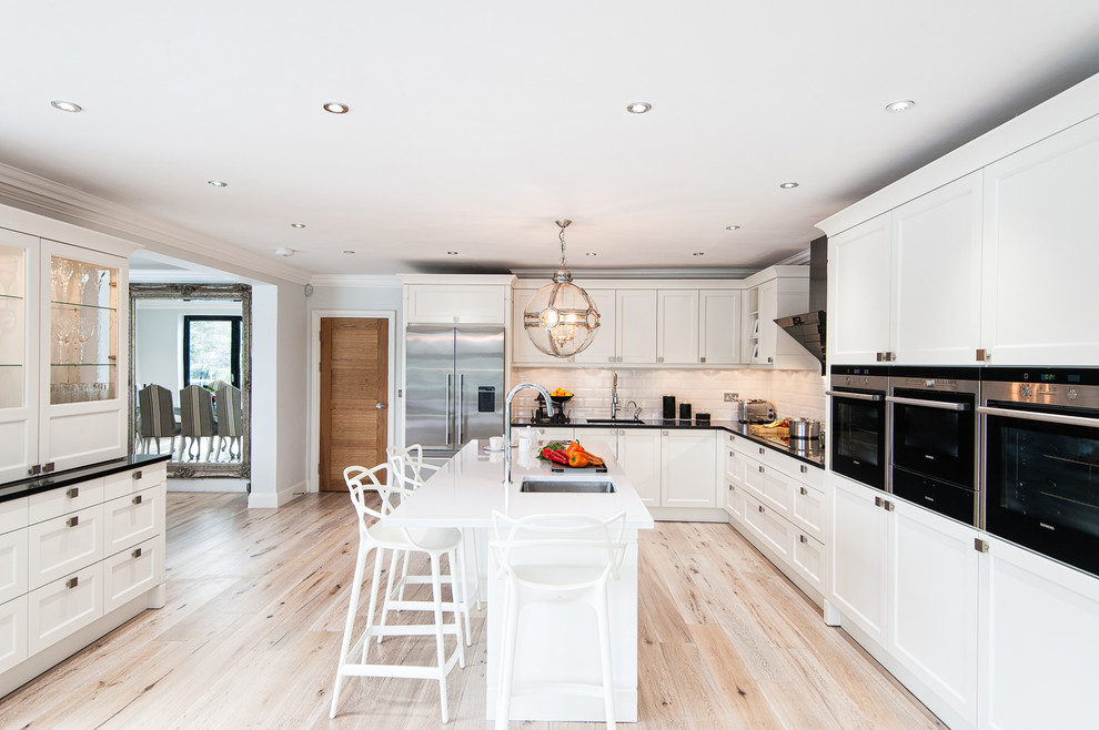Modern inredning av ett stort kök, med en nedsänkt diskho, luckor med infälld panel, vita skåp, bänkskiva i kvartsit, vitt stänkskydd, stänkskydd i tunnelbanekakel, rostfria vitvaror, ljust trägolv och en köksö