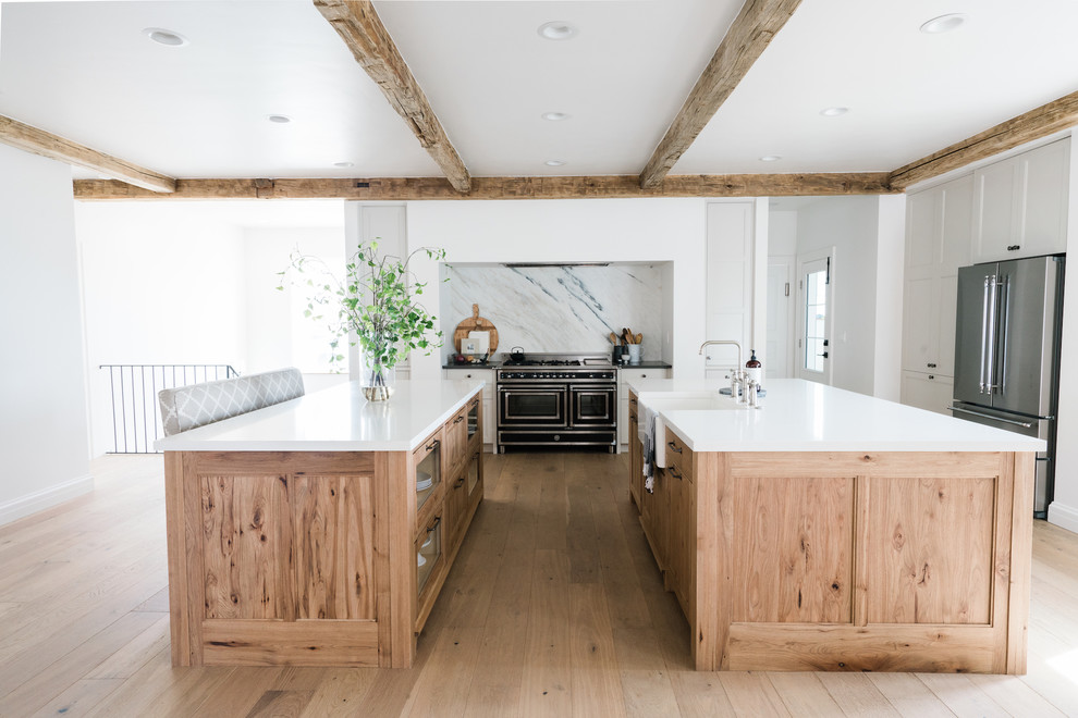 This is an example of a country kitchen in Los Angeles with a belfast sink, glass-front cabinets, light wood cabinets, grey splashback, stone slab splashback, stainless steel appliances, light hardwood flooring, multiple islands, brown floors and white worktops.