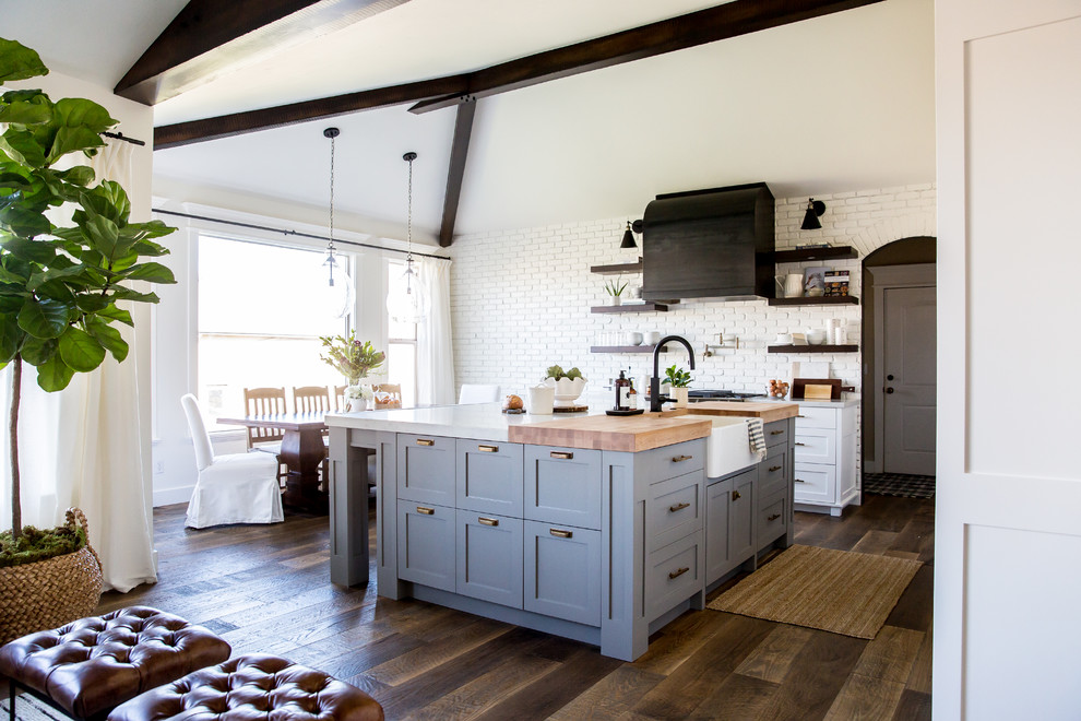 Bild på ett mellanstort lantligt kök, med en rustik diskho, skåp i shakerstil, vita skåp, bänkskiva i kvarts, vitt stänkskydd, stänkskydd i tegel, svarta vitvaror, mellanmörkt trägolv och en köksö