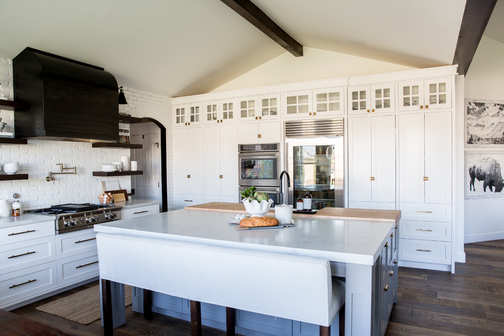 Example of a mid-sized cottage l-shaped medium tone wood floor eat-in kitchen design in Salt Lake City with a farmhouse sink, shaker cabinets, white cabinets, quartz countertops, white backsplash, brick backsplash, stainless steel appliances and an island