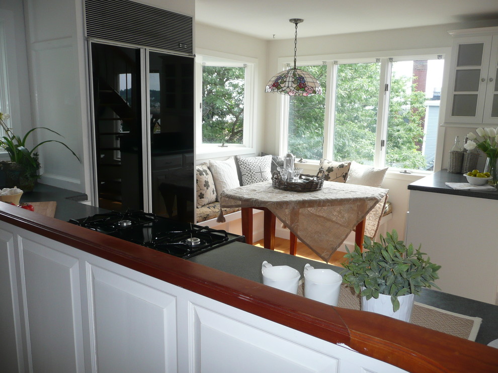 Photo of a medium sized coastal l-shaped kitchen/diner in Boston with a built-in sink, glass-front cabinets, white cabinets, laminate countertops, white splashback, black appliances, light hardwood flooring and a breakfast bar.