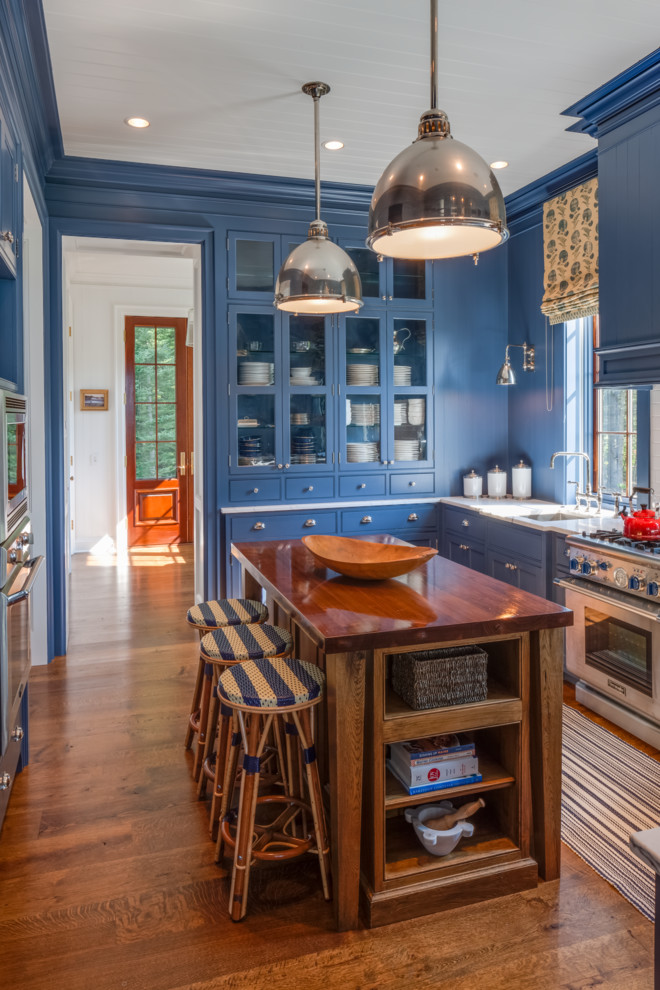 Example of a beach style l-shaped medium tone wood floor enclosed kitchen design in Portland Maine with a single-bowl sink, glass-front cabinets, blue cabinets, wood countertops, stainless steel appliances and an island