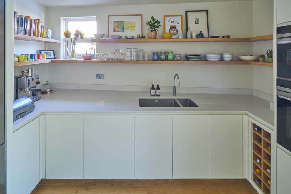 This is an example of a medium sized contemporary u-shaped kitchen/diner in London with an integrated sink, flat-panel cabinets, white cabinets, white splashback, stainless steel appliances, medium hardwood flooring and no island.