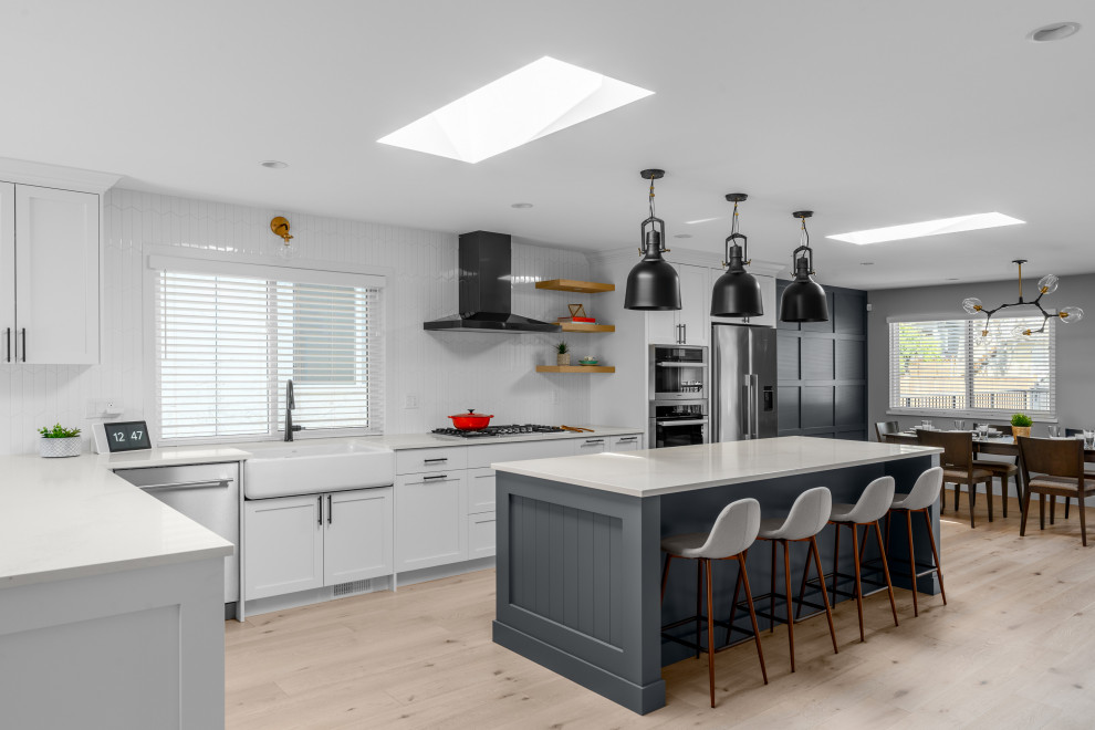 Design ideas for a large classic l-shaped kitchen/diner in Vancouver with a belfast sink, shaker cabinets, white cabinets, engineered stone countertops, white splashback, porcelain splashback, stainless steel appliances, light hardwood flooring, an island, brown floors and white worktops.