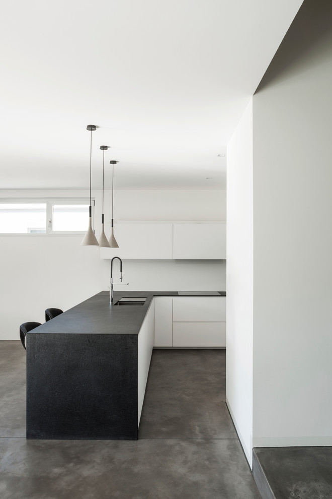 На фото: угловая кухня в стиле модернизм с врезной мойкой, плоскими фасадами, бетонным полом, полуостровом и черно-белыми фасадами