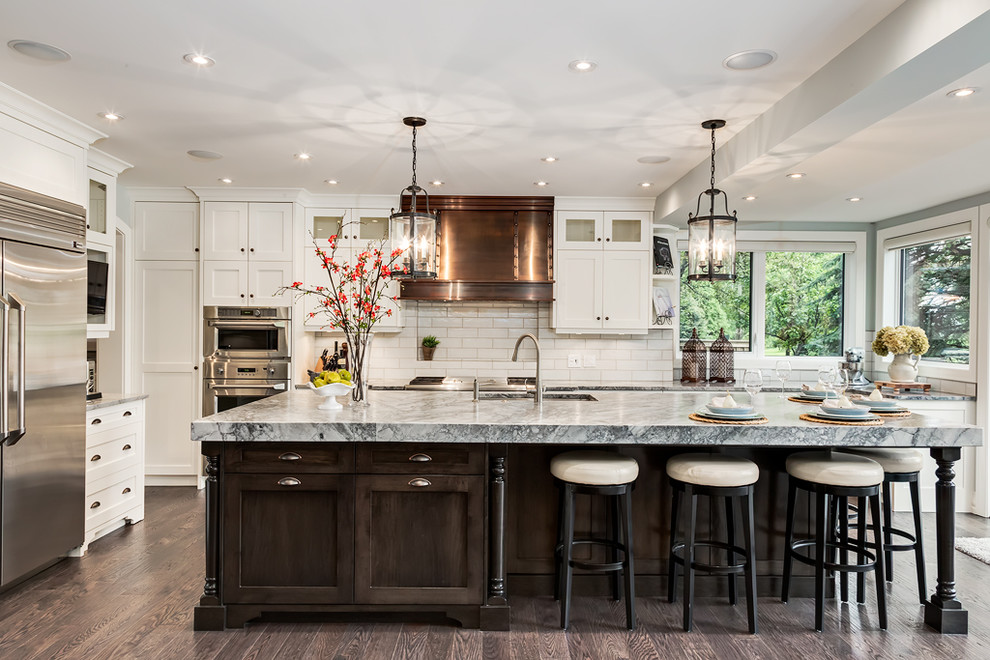 Medium sized traditional l-shaped enclosed kitchen in Calgary with a submerged sink, shaker cabinets, white cabinets, white splashback, metro tiled splashback, stainless steel appliances, dark hardwood flooring, an island and brown floors.